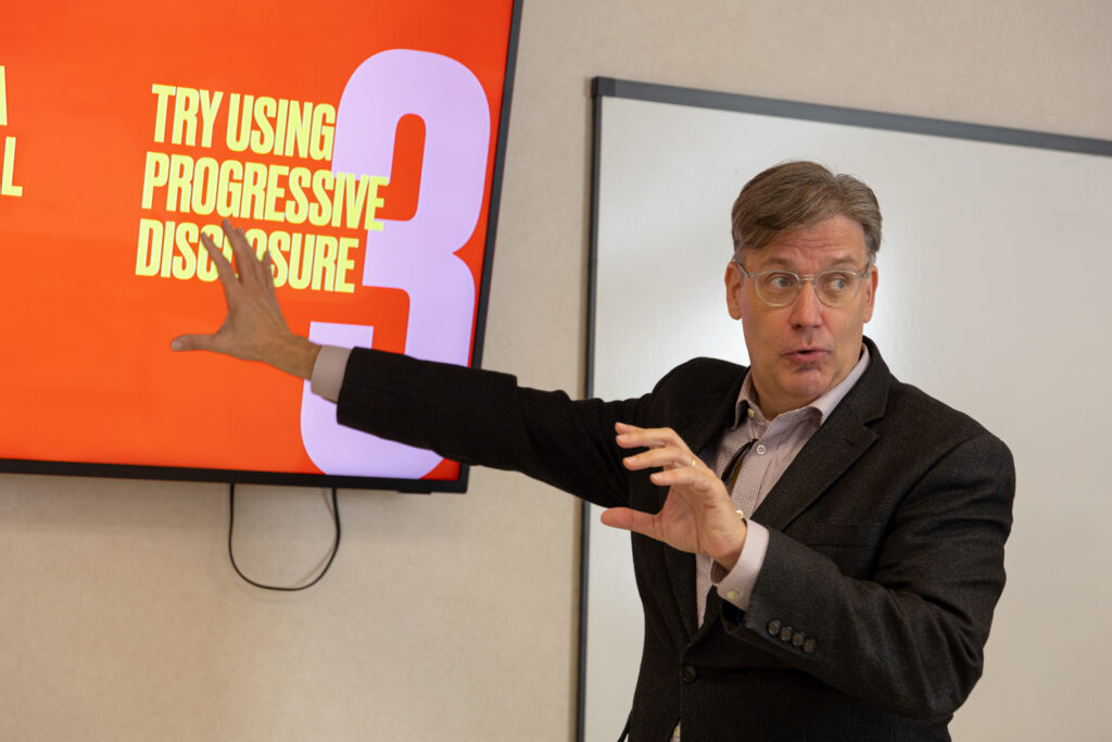A man in a suit gestures toward a large screen displaying the text “TRY USING PROGRESSIVE DISCLOSURE 3” in bold yellow and white letters on a red background—a modern approach to digital communications. A whiteboard is visible in the background.