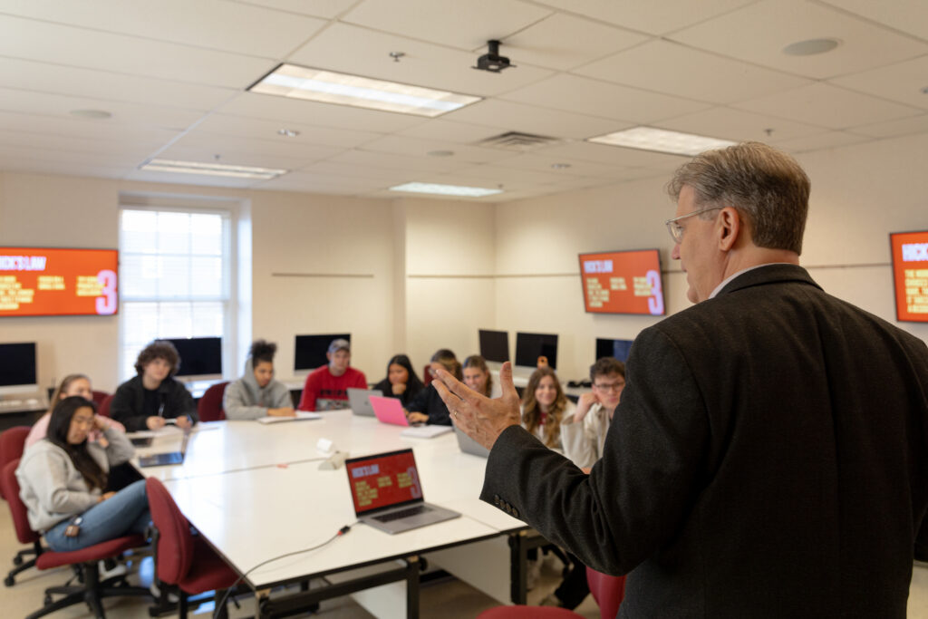 A man in a suit stands at the front of a well-lit classroom, engaging students in an Interactive Digital Media Communications session. Laptops and multiple monitors displaying 