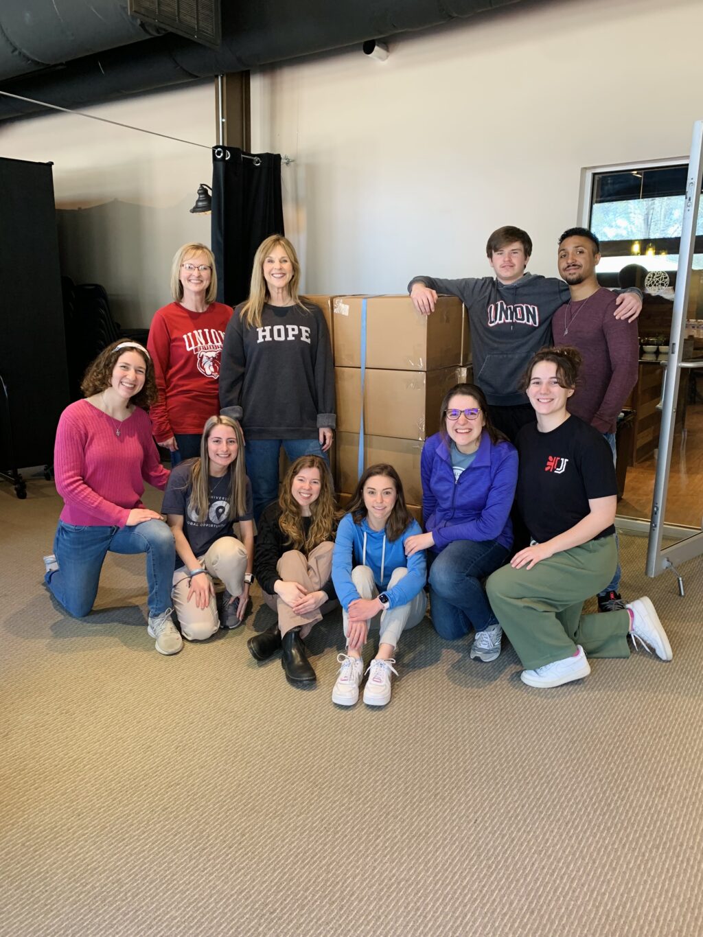 A group of eleven people from a Christian Ministry pose and smile together in front of stacked cardboard boxes indoors. Some are sitting on the floor, while others stand behind them. Most are wearing casual clothes and sweatshirts.