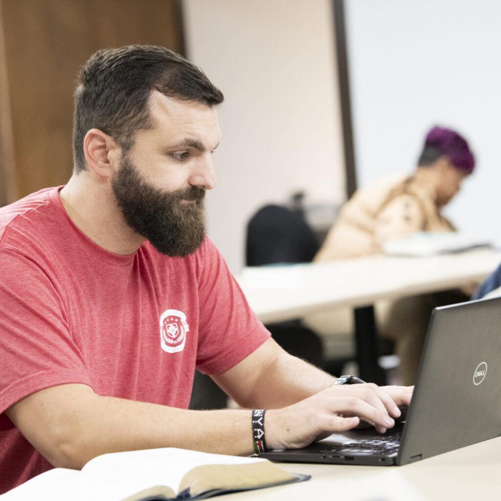 A man with a beard wearing a red t-shirt types on a Dell laptop at a desk, possibly studying for his Associate of General Studies degree. An open book is in front of him. In the background, another person looks down at papers.