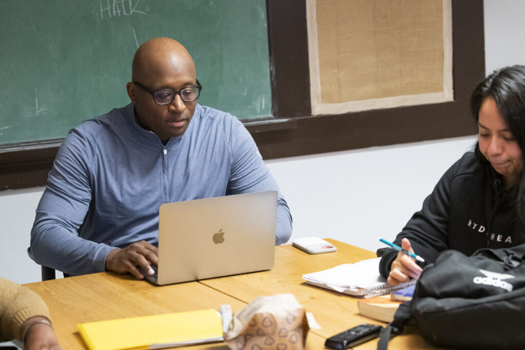 A man in glasses works on a laptop at a wooden table, while a woman next to him writes in a notebook—perhaps both studying for their Associate of Science or Professional Studies degree. Books, papers, and a backpack fill the table near a green chalkboard.