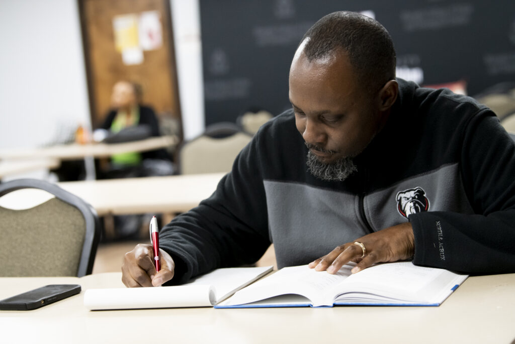 A man sits at a table in a classroom, engaged in Urban Theological Studies as he writes in a notebook with a pen while reading an open textbook. Other empty chairs and a person in the background are visible.