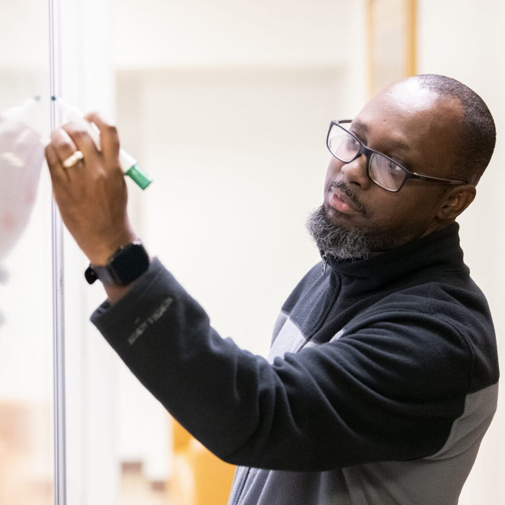 A man wearing glasses and a black and gray jacket writes on a whiteboard with a green marker in a brightly lit room during an Urban Theological Studies session.