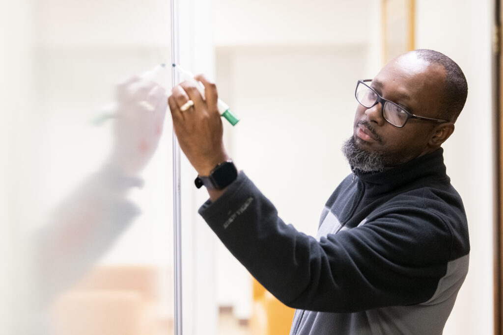 A man wearing glasses and a black jacket writes on a whiteboard with a green marker in a brightly lit room, possibly discussing topics related to an Associate of Science or Professional Studies program.