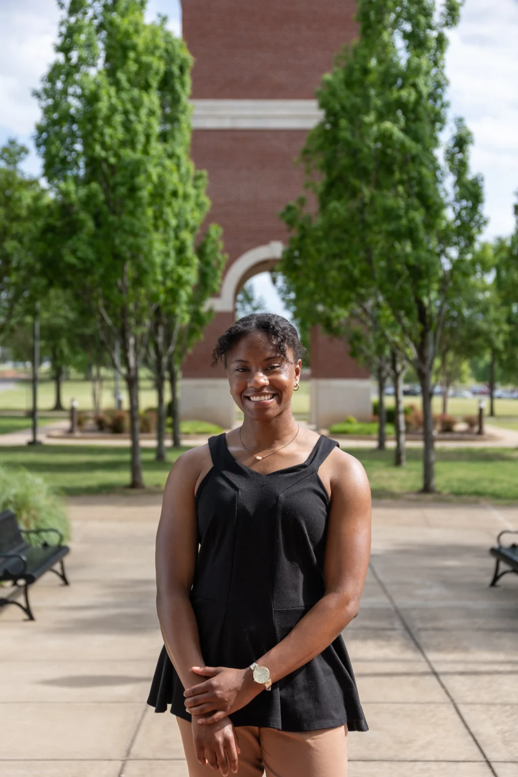 Makayla Madison, in a black sleeveless top, stands outdoors on a paved walkway, smiling at the camera. Behind her are green trees, benches, and a brick structure with an archway.