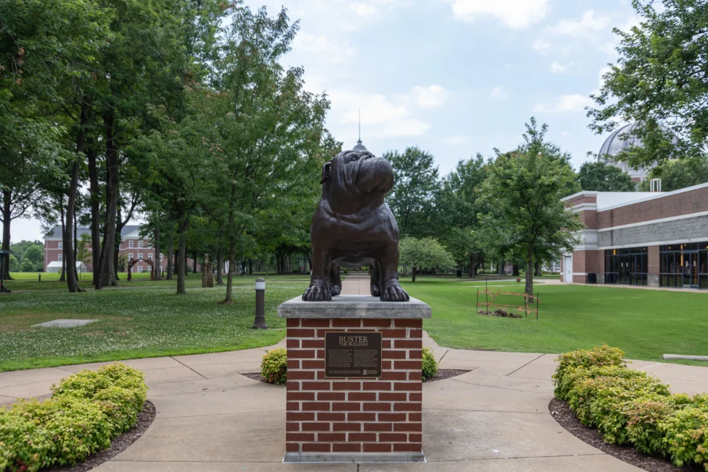 A bronze bulldog statue stands on a brick pedestal with a plaque, surrounded by a landscaped path and green lawn, with trees and campus buildings in the background.