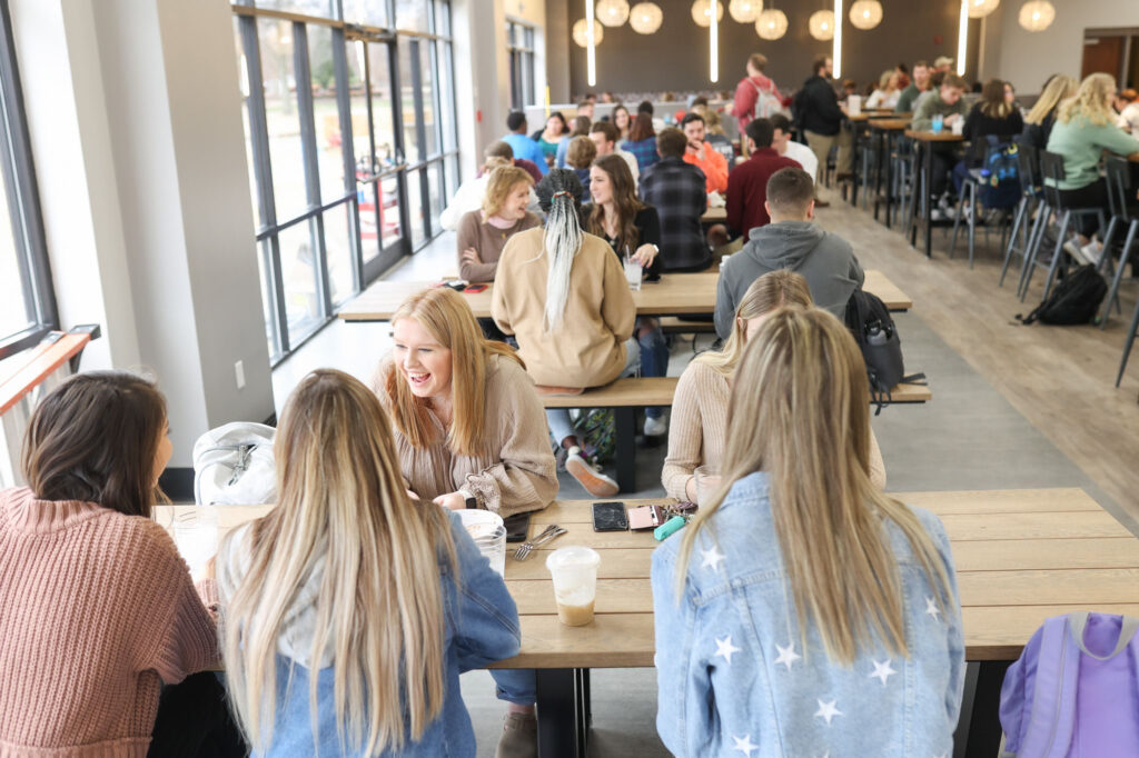 A group of young people sit at long wooden tables in a busy, modern cafe with large windows. Some are talking and laughing, while others focus on their drinks or conversations. Round light fixtures hang from the ceiling.