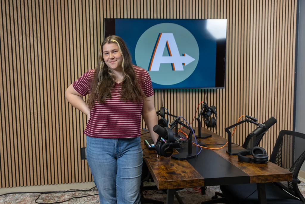 Norah Taylor, with long hair, wears a red striped shirt and jeans as she stands by a wooden podcast table equipped with microphones, headphones, and cables. A screen behind her displays a stylized A logo.