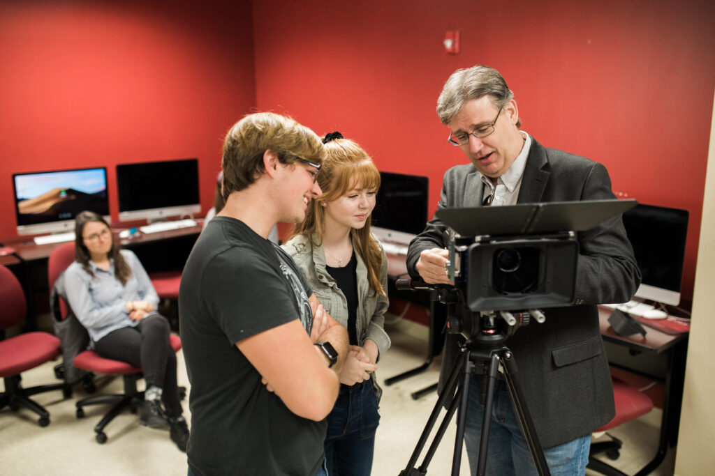 A man in a suit jacket demonstrates a camera setup on a tripod to two students in a Digital Media Communications lab with red walls, while another person sits at a computer in the background.