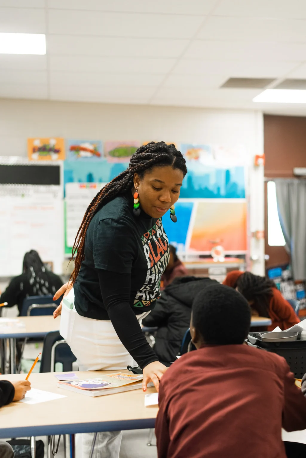 A teacher wearing a Black History Month t-shirt leans over to help a student at a desk in a classroom. Other students are seated and working, with educational posters on the classroom walls.
