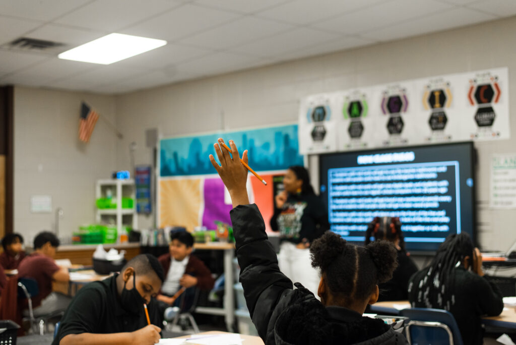 A student with a raised hand sits at a desk in an EdS classroom. Other students are writing, while an instructor stands at the front near a large screen displaying text—emphasizing leadership in school reform. Classroom decor and an American flag are visible.