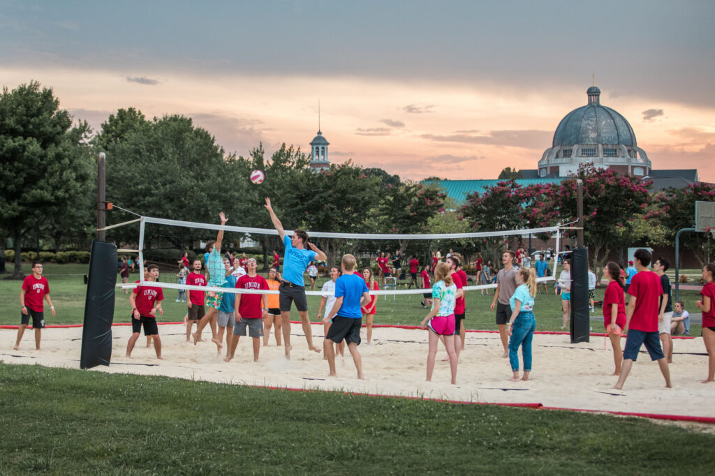 A group of people playing a game of volleyball on a sand court in a park during sunset, with trees and domed buildings visible in the background. Spectators stand around the court.