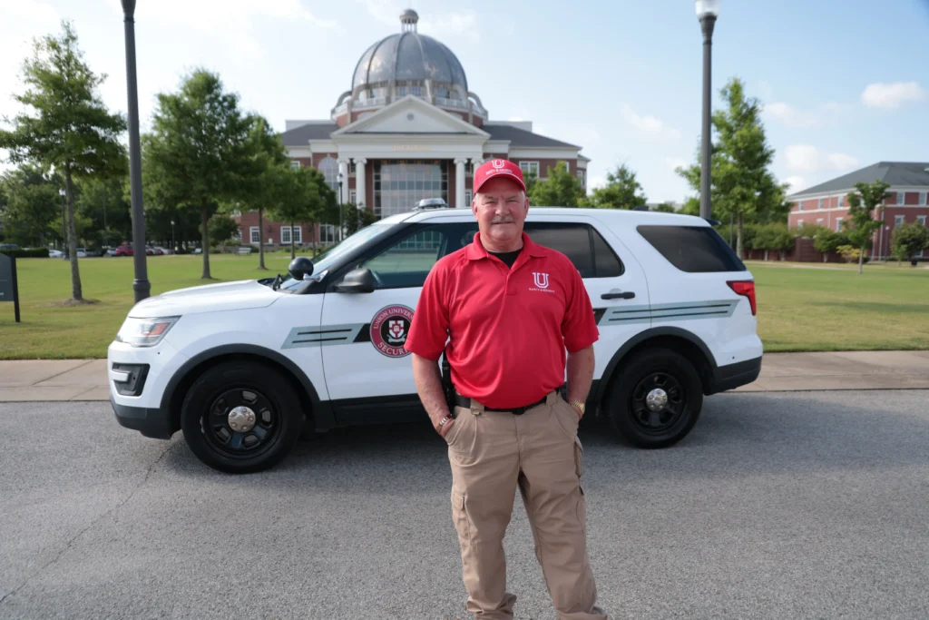 A man in a red polo and khaki pants stands before a white university police SUV, highlighting the active Campus Life scene with a domed building and trees providing a classic collegiate backdrop.