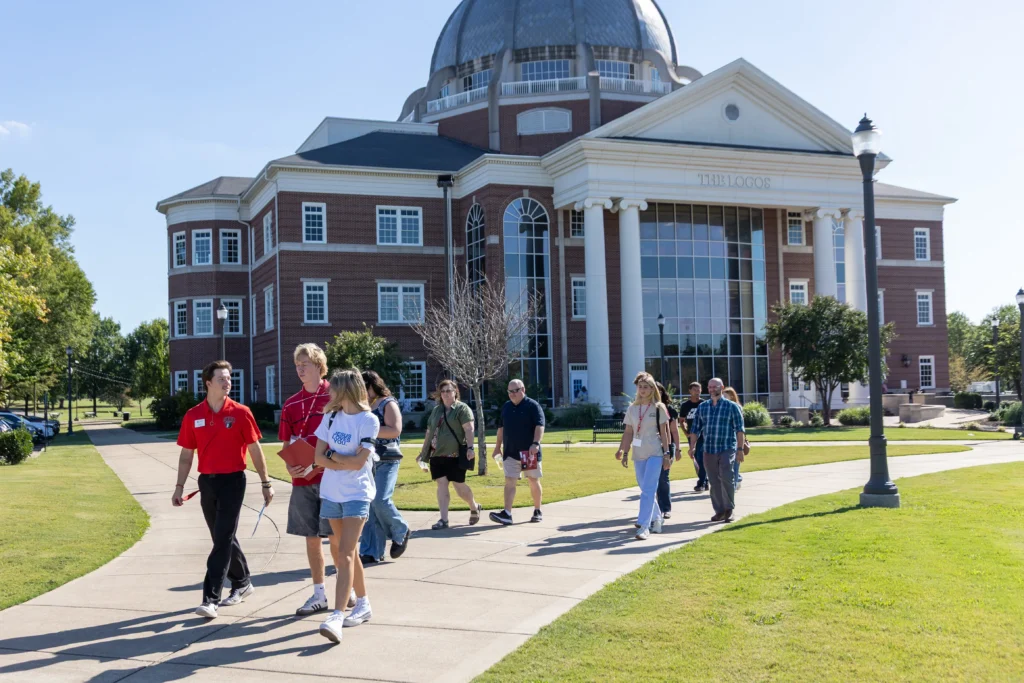 A group of people walks along a sidewalk in front of a large, multi-story brick building with tall white columns and a domed roof on a sunny day.