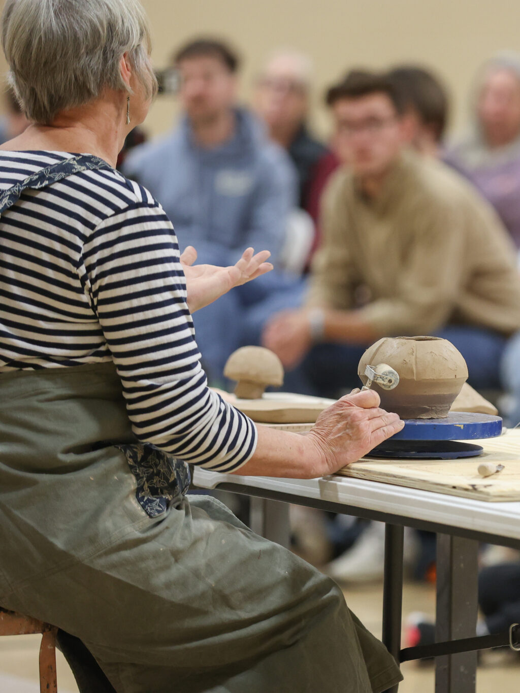 A person with short gray hair, wearing a striped shirt and apron, gives a pottery demonstration to a seated audience. They gesture with one hand while a clay piece sits on a spinning wheel in front of them.
