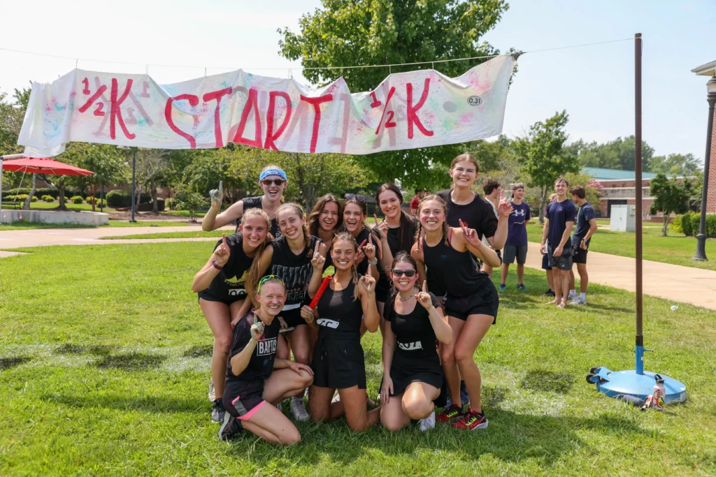 A group of eleven smiling people pose in front of a handmade 1/2K START 1/2K sign outdoors on a sunny day, capturing the energy of Student Life. Most wear black athletic clothing, and several are holding up one finger.