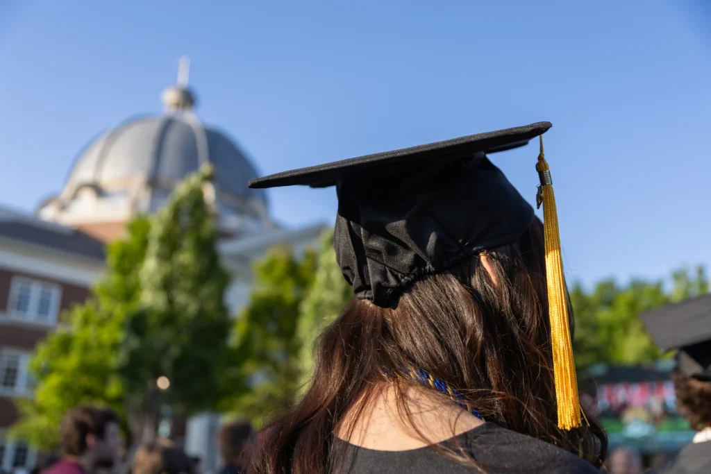 A person with long brown hair wearing a graduation cap and tassel stands outdoors, with a blurred building featuring a dome and trees in the background.