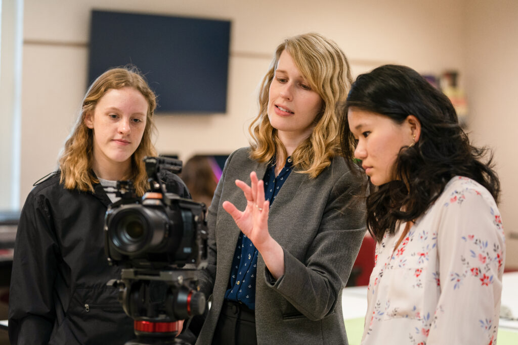 Three women stand together in an office setting, looking at a video camera on a tripod. One woman gestures while explaining something related to Digital Media Studies as the other two listen attentively. A monitor is visible in the background.