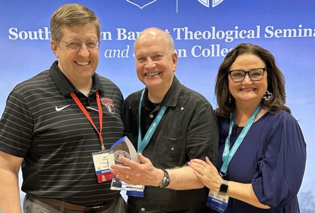 Three adults stand together smiling in front of a Southern Baptist Theological Seminary banner. The man in the center proudly holds the Dodd Award, while the others—representing Union and Akin—stand beside him, touching his arm.