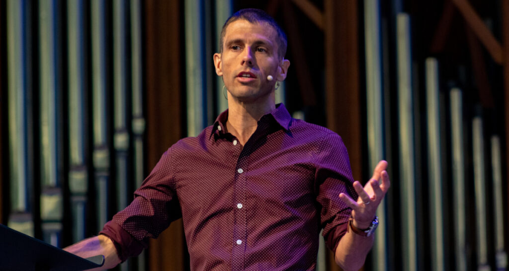 A person wearing a maroon, patterned shirt speaks on stage with a headset microphone, gesturing with one hand during the Powell Missions Lecture on March 10. Large metallic pipes are visible in the background.