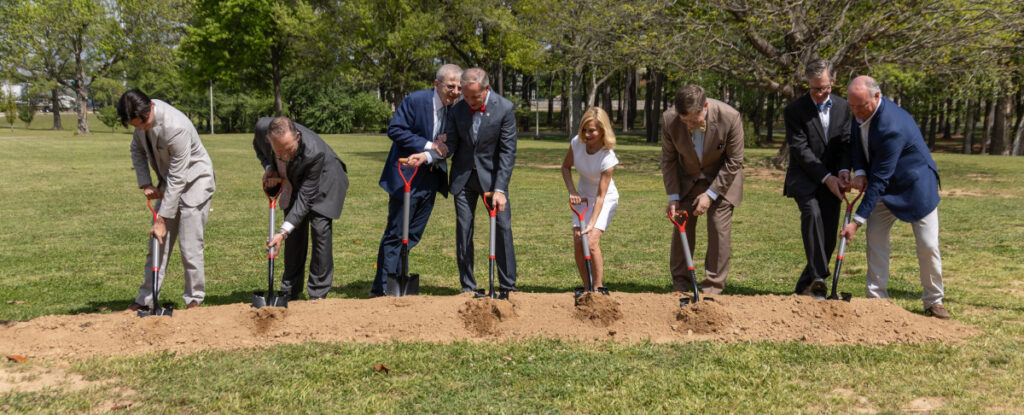 Eight people in business attire stand in a row outdoors on grass, using shovels to break ground for Union’s new $30 million chapel. Trees and green space are visible in the background, highlighting the ceremonial event.