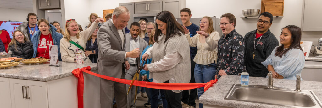 A man and a woman cut a red ribbon with large scissors in their new home’s kitchen as part of the Union EDGE program, while a group of people cheer and smile. Food platters and drinks are set out on the countertops.