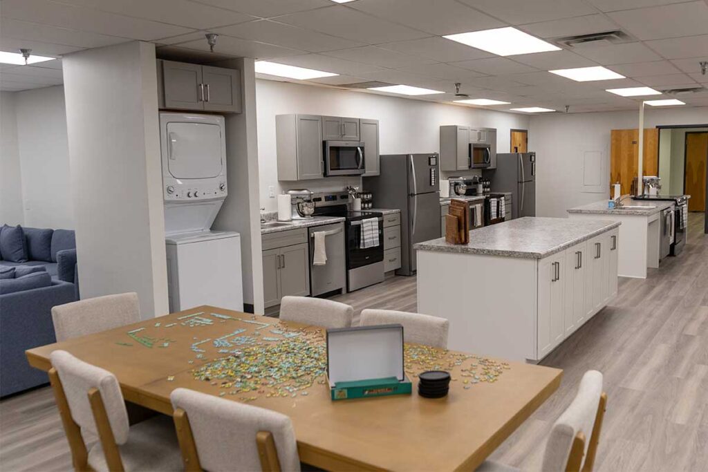 A modern communal kitchen and dining area with gray cabinets, stainless steel appliances, a kitchen island, and a wooden table with chairs. A partially completed jigsaw puzzle and box are on the table.