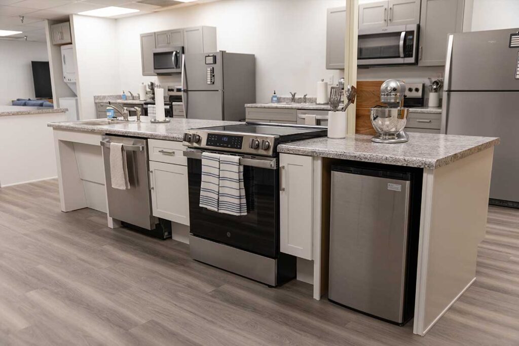 A modern kitchen with gray cabinets, stainless steel appliances, a stove with towels hanging on the handle, a stand mixer, a dishwasher, a refrigerator, and a microwave on the countertops. The floor is wood-patterned.