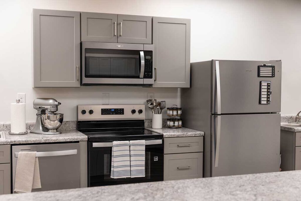 Modern kitchen with gray cabinets, stainless steel appliances including a microwave, oven, and refrigerator, a stand mixer, spice jars, a paper towel holder, and dish towels hanging on the drawers and oven handle.