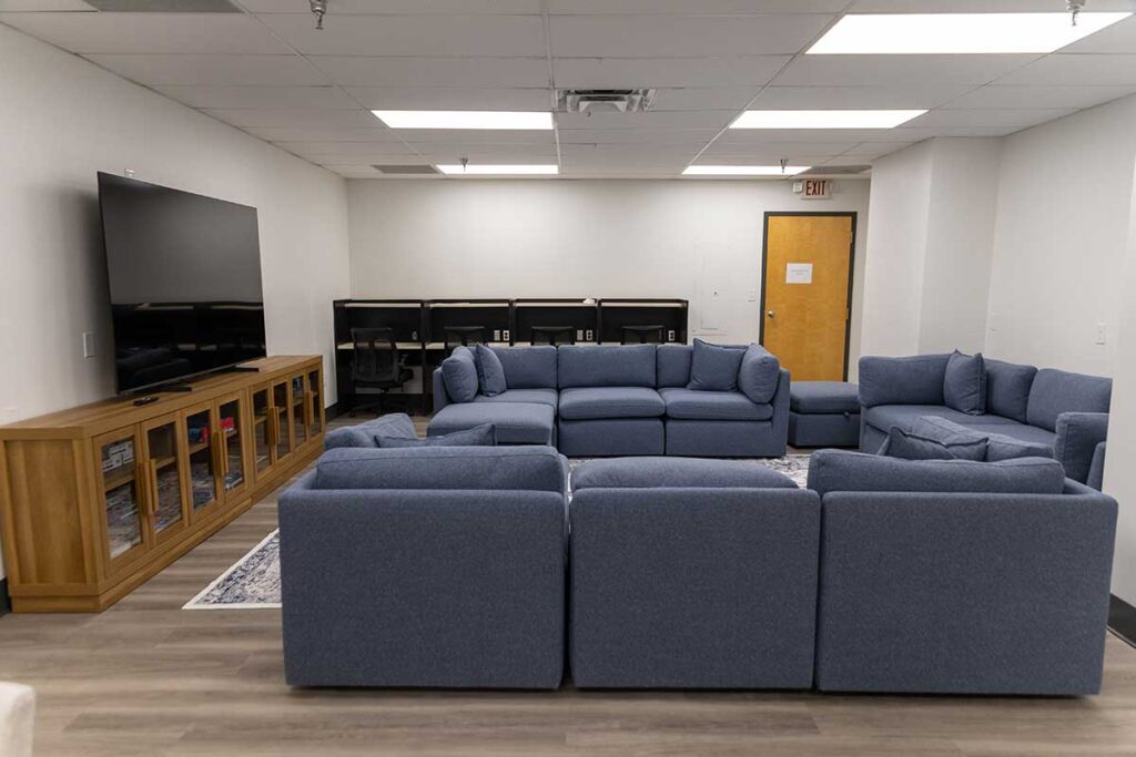 A large room with blue sectional sofas arranged around a rug, a wooden TV stand with a flat-screen TV, and desks with chairs along the back wall near a closed wooden door. Ceiling has fluorescent lights.
