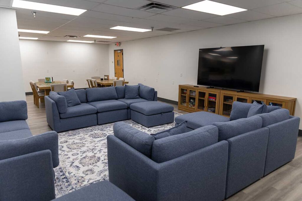 A spacious lounge with two blue sectional sofas arranged around a patterned rug, facing a large flat-screen TV on a wooden console; tables and chairs are visible in the background.