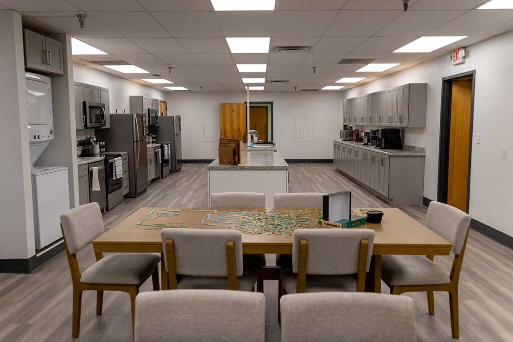 A spacious kitchen and dining area with light wood flooring, gray cabinets, stainless steel appliances, and a table with six chairs. A partially completed jigsaw puzzle and open puzzle box are on the table.