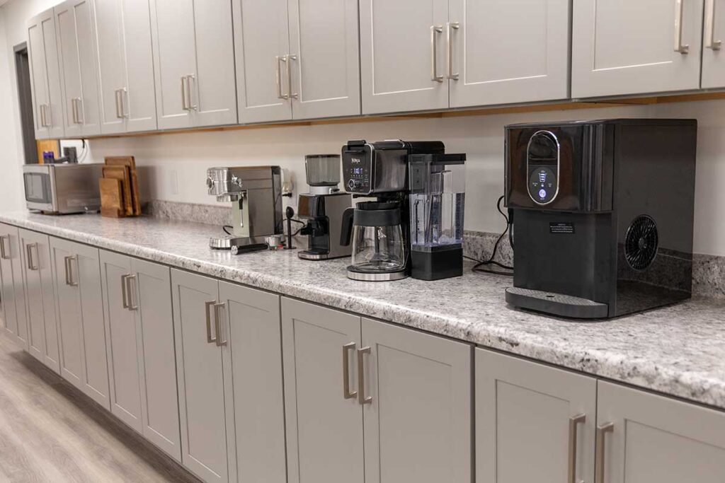 A kitchen counter with grey cabinets, featuring a microwave, espresso machine, drip coffee maker, and a modern water or ice dispenser. The appliances are positioned in a row on a speckled countertop.