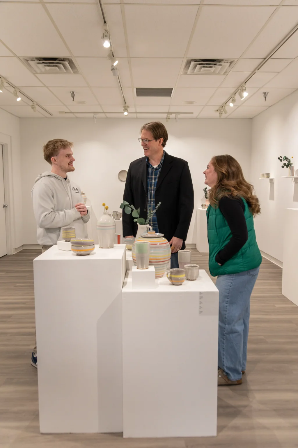 Three people stand around a white display pedestal with ceramic pots and cups in a gallery. The room has white walls, wooden flooring, and shelves with small ceramic pieces. The group is smiling and talking.