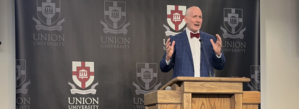 A man in a blue suit and red bow tie speaks at a wooden podium in front of a backdrop with the Union University logo, reflecting the institution's strong Baptist heritage and evangelical identity.