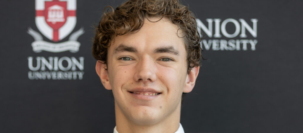 A young man with curly brown hair smiles at the camera in front of a backdrop with the Union University logo and name. Gutierrez is wearing a white shirt and dark tie, celebrating earning Union’s top academic scholarship.