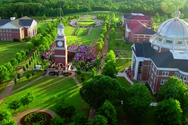 Aerial view of a university campus with brick buildings, a central clock tower, landscaped lawns, trees, flowerbeds, and a large group of people gathered outdoors, possibly for a commencement ceremony.