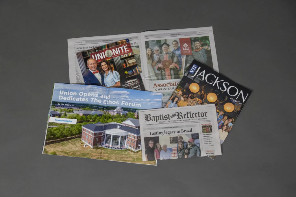 Five publications are arranged on a gray surface, including newspapers, newsletters, and magazines featuring headlines, photos of people, and a building on their covers.