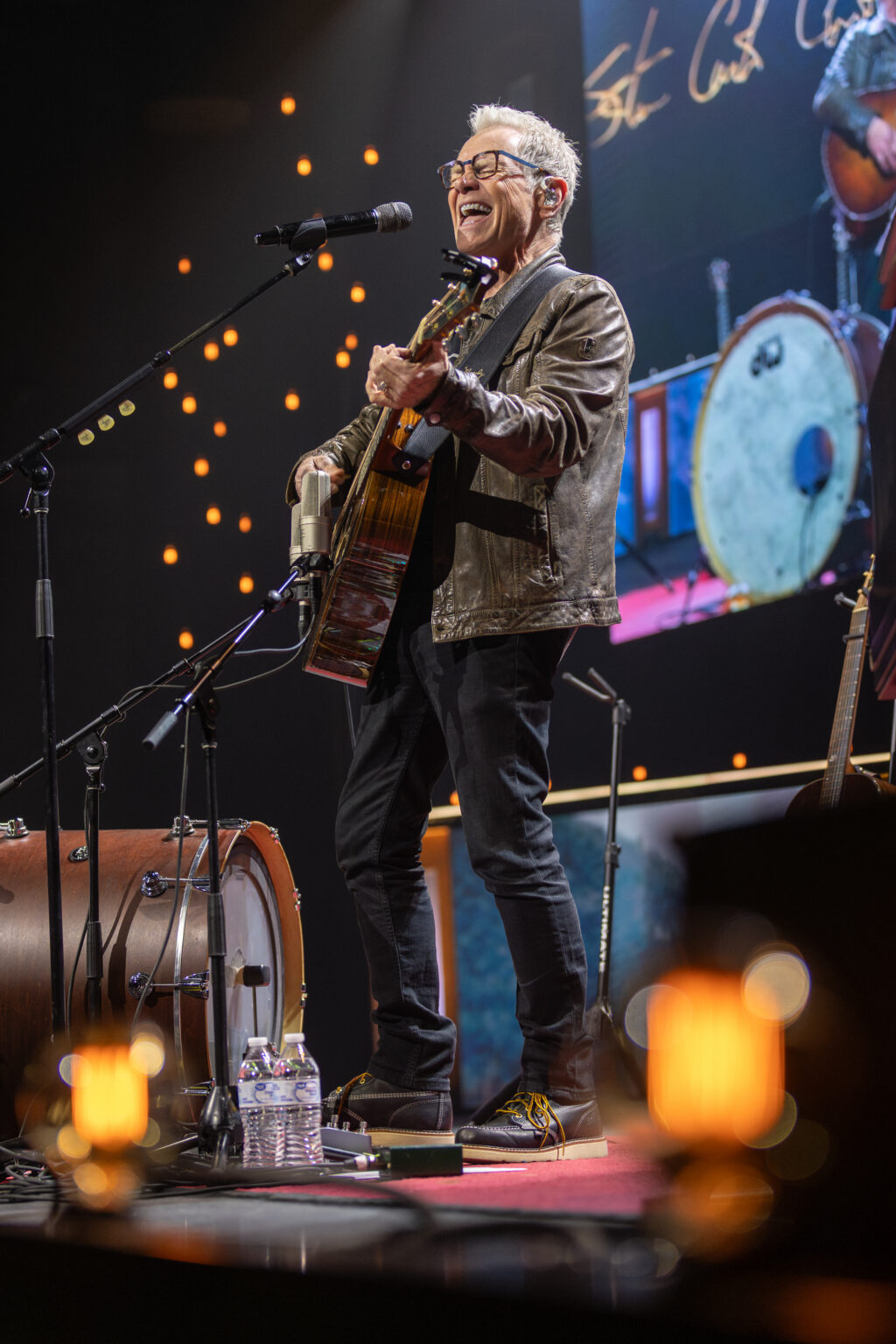 A man wearing glasses, a brown leather jacket, black pants, and sneakers plays an acoustic guitar and sings into a microphone on stage, with a drum and water bottles nearby and stage lights in the background.
