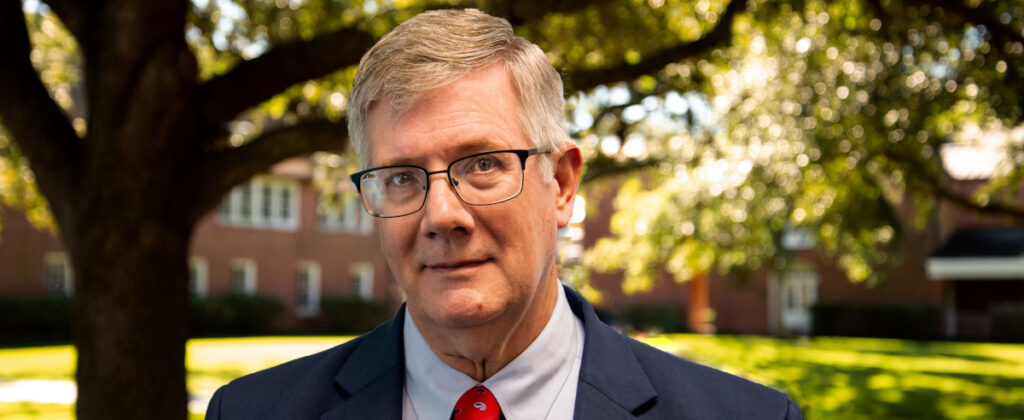 A man with gray hair and glasses, wearing a navy suit, light blue shirt, and red tie, stands outdoors in front of a tree and a brick building, attending the Ryan Center Bible Conference on March 20-21.