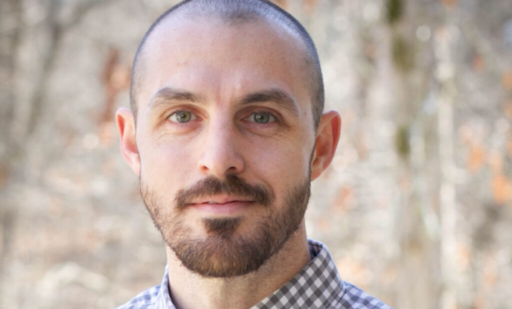 A man with a shaved head and trimmed beard, wearing a checkered shirt, stands outdoors in front of a blurred background of trees, possibly attending the Union or UAB infectious disease James Scholar-in-Residence Lectures.