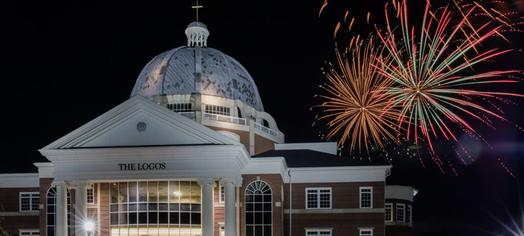 A brick building with a white dome labeled The Logos is seen at night, with colorful fireworks lighting up the dark sky in the background.