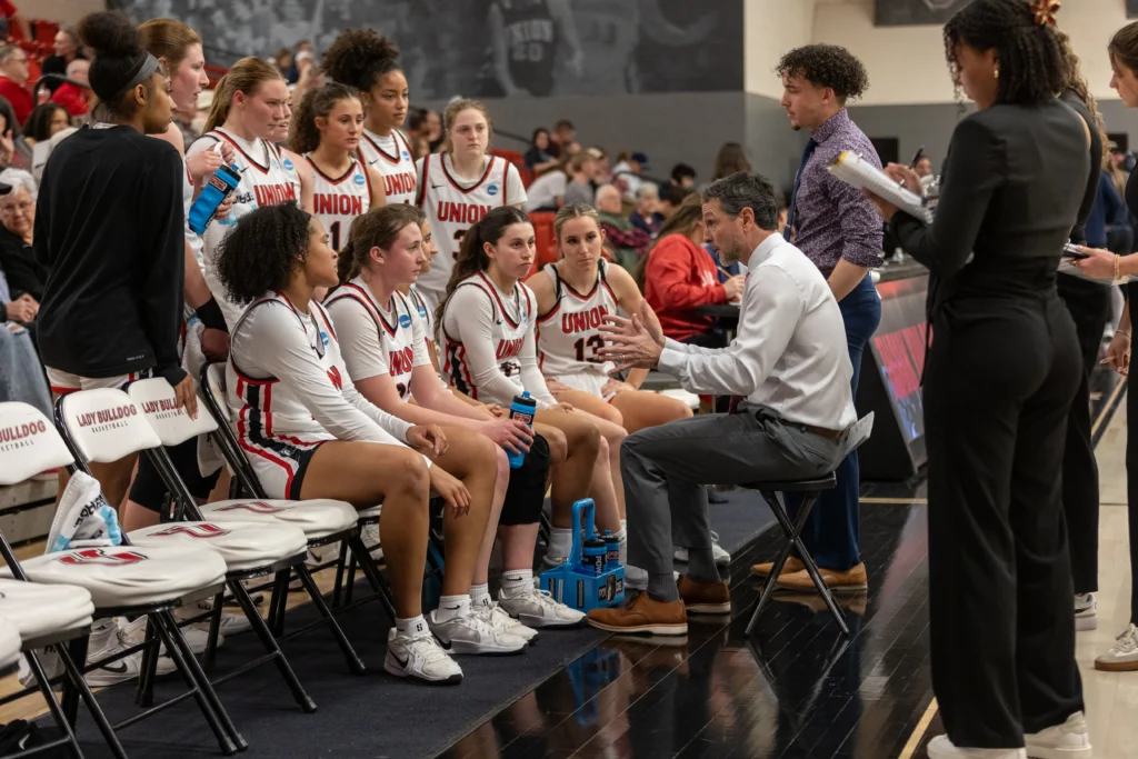 A womens basketball team in Union uniforms sits on a bench during a timeout, listening to their coach, who is kneeling and talking. Assistant coaches and staff stand nearby. The gym is filled with spectators.