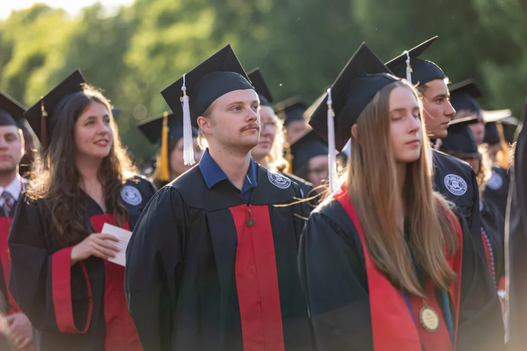 A group of college graduates wearing caps and gowns stand outdoors during a graduation ceremony. Some are looking forward, and sunlight shines on their faces. Trees are visible in the background.