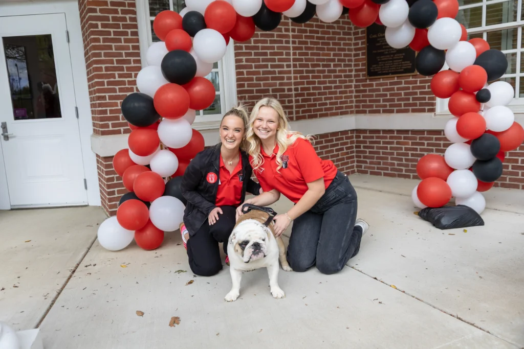 Two women kneel on a sidewalk next to a bulldog in front of a red, white, and black balloon arch, with a brick building and white door in the background.