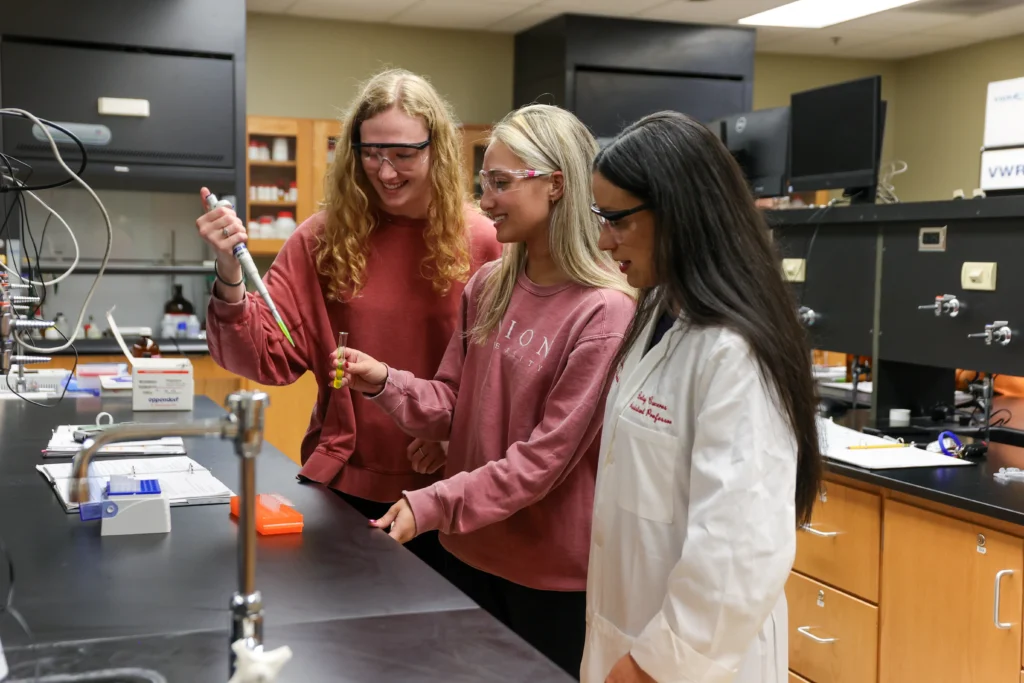 Three women in safety glasses work together at a lab bench. One uses a pipette while the others observe. They are surrounded by laboratory equipment and cabinets in a science classroom setting.