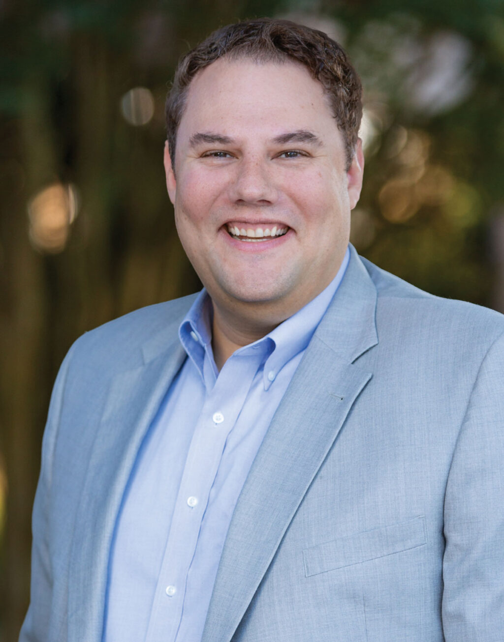 A man with short brown hair, wearing a light blue dress shirt and light gray blazer, smiles at the camera. The background is blurred greenery, creating a professional yet approachable atmosphere—an image well-suited for Gray Coyner of Next Generation Investing.