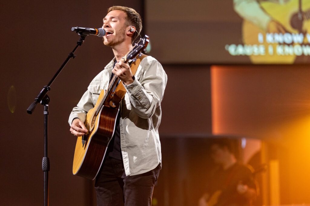 A man singing into a microphone and playing an acoustic guitar on stage. He wears a light jacket and has in-ear monitors. There is another musician and a projected screen with partial song lyrics in the background.