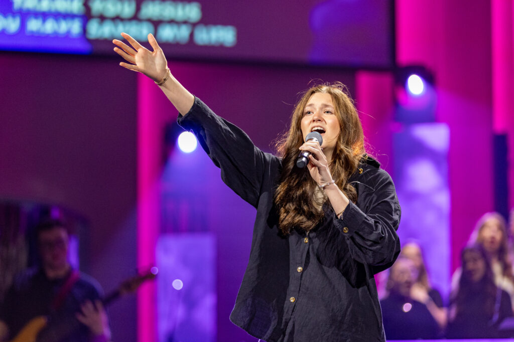 A woman sings into a microphone with one arm raised on a stage, lit by purple and blue lights. People and musical instruments are visible in the background.