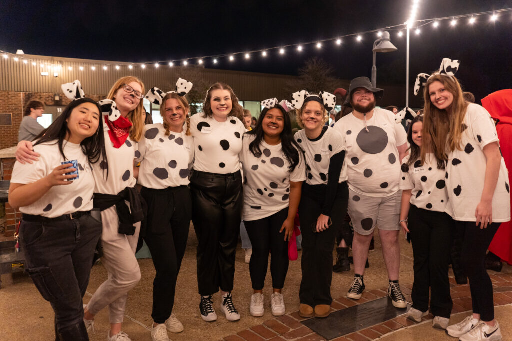 A group of people dressed as dalmatians pose for a photo outdoors at night. They wear white shirts with black spots and dog ear headbands. String lights hang above them, and other people are visible in the background.